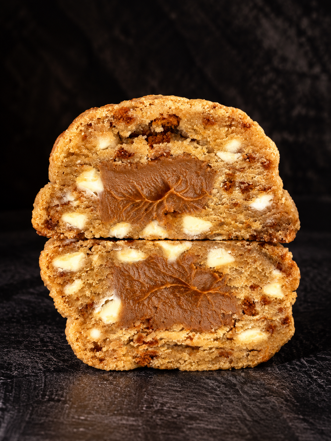 Two halves of a large chocolate chip cookie stacked vertically, showing a thick biscoff spread filling and white chocolate chips, set against a dark background.