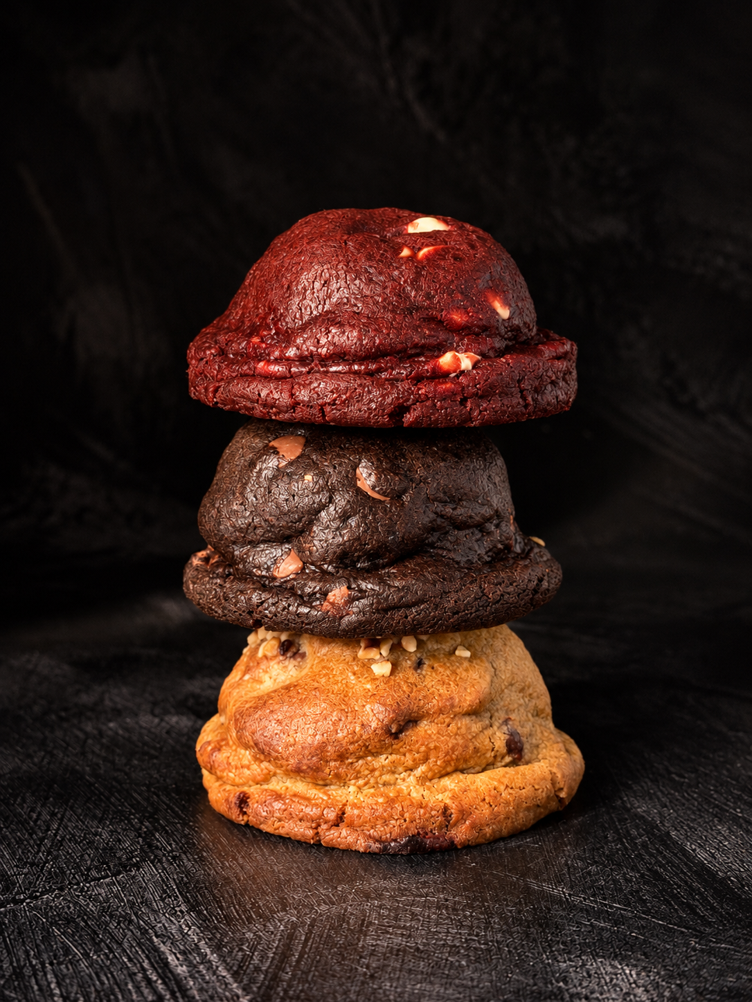 Stack of thick handmade Black Bird Bakers cookies in red velvet, chocolate and pistachio flavours on a dark textured background